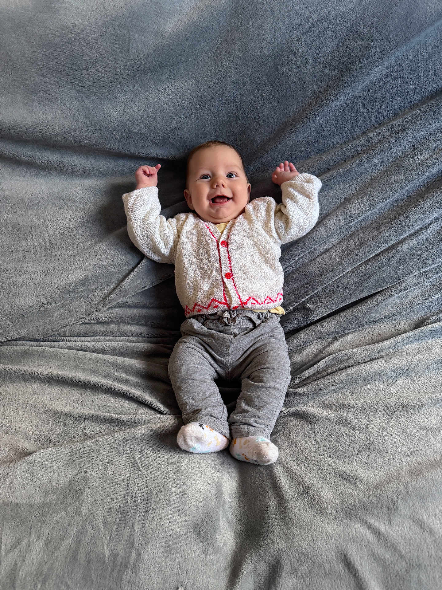 Judith participe au concours pour gagner de l'argent avec cette photo : baby, smiling, infant, blanket, gray_blanket, cardigan, red_buttons, gray_pants, white_socks, happy, cute, lying_down, raised_arms, soft_texture, indoors, child, portrait, cozy, warm_clothing, small_feet