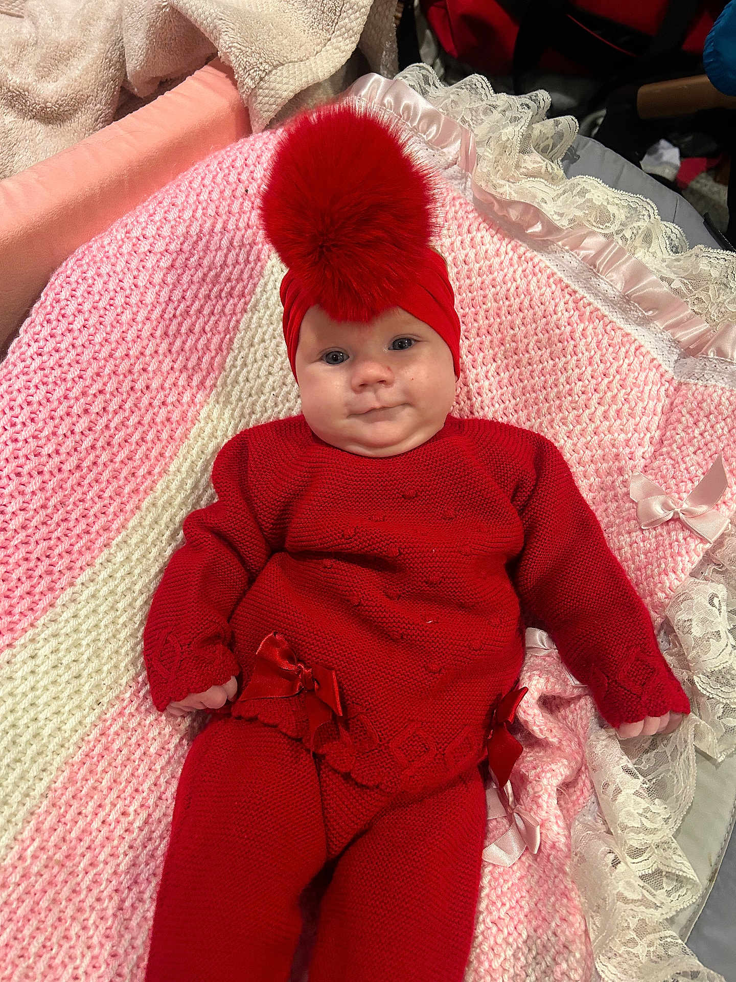 Dolcie is registered to the contest to win money with this photo: baby, blanket, bow, child, cozy, cute, hat, indoors, infant, knitted_outfit, lace, lying_down, pink, pom_pom, portrait, red_clothing, smile, soft_texture, warm_clothing, white