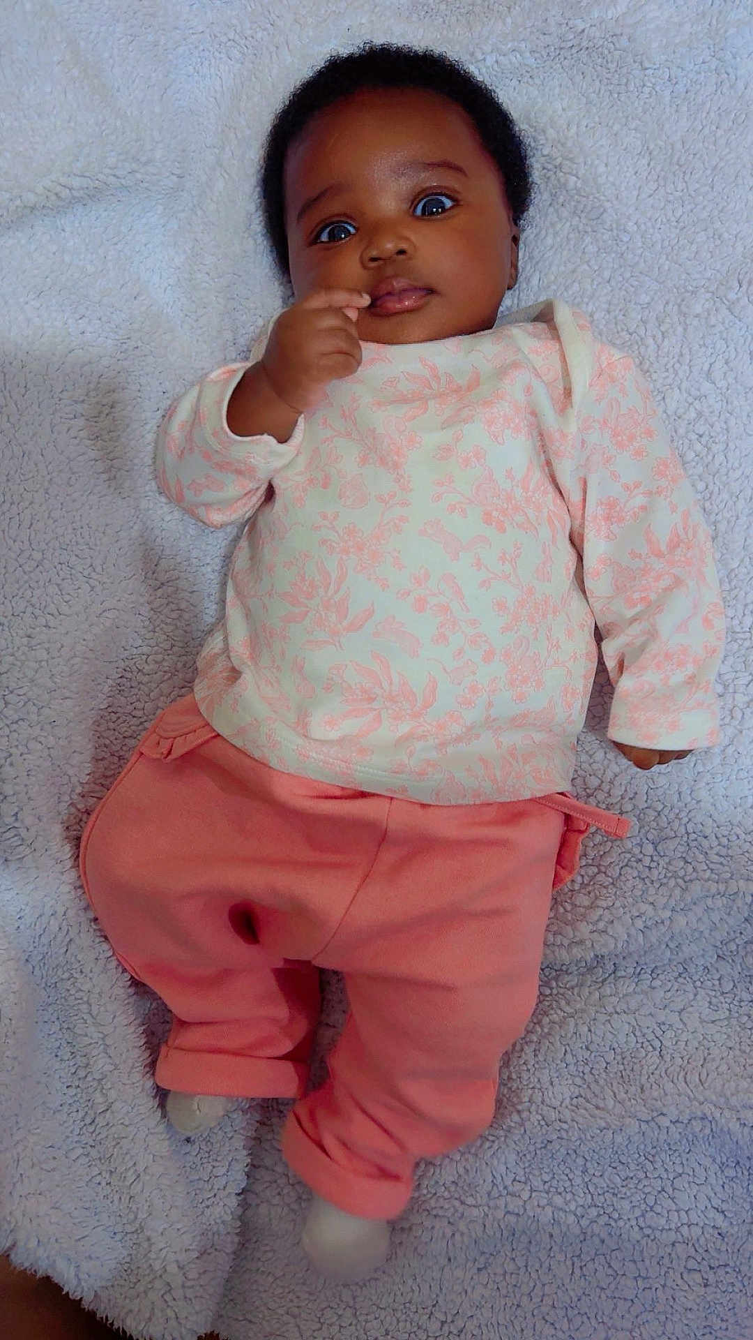 Jesulayomi is registered to the contest to win money with this photo: baby, infant, child, pink_pants, floral_top, blanket, lying_down, curious, wide_eyes, hand_near_mouth, soft_texture, indoors, cute, portrait, closeup, skin, person, cozy, sleepwear, young