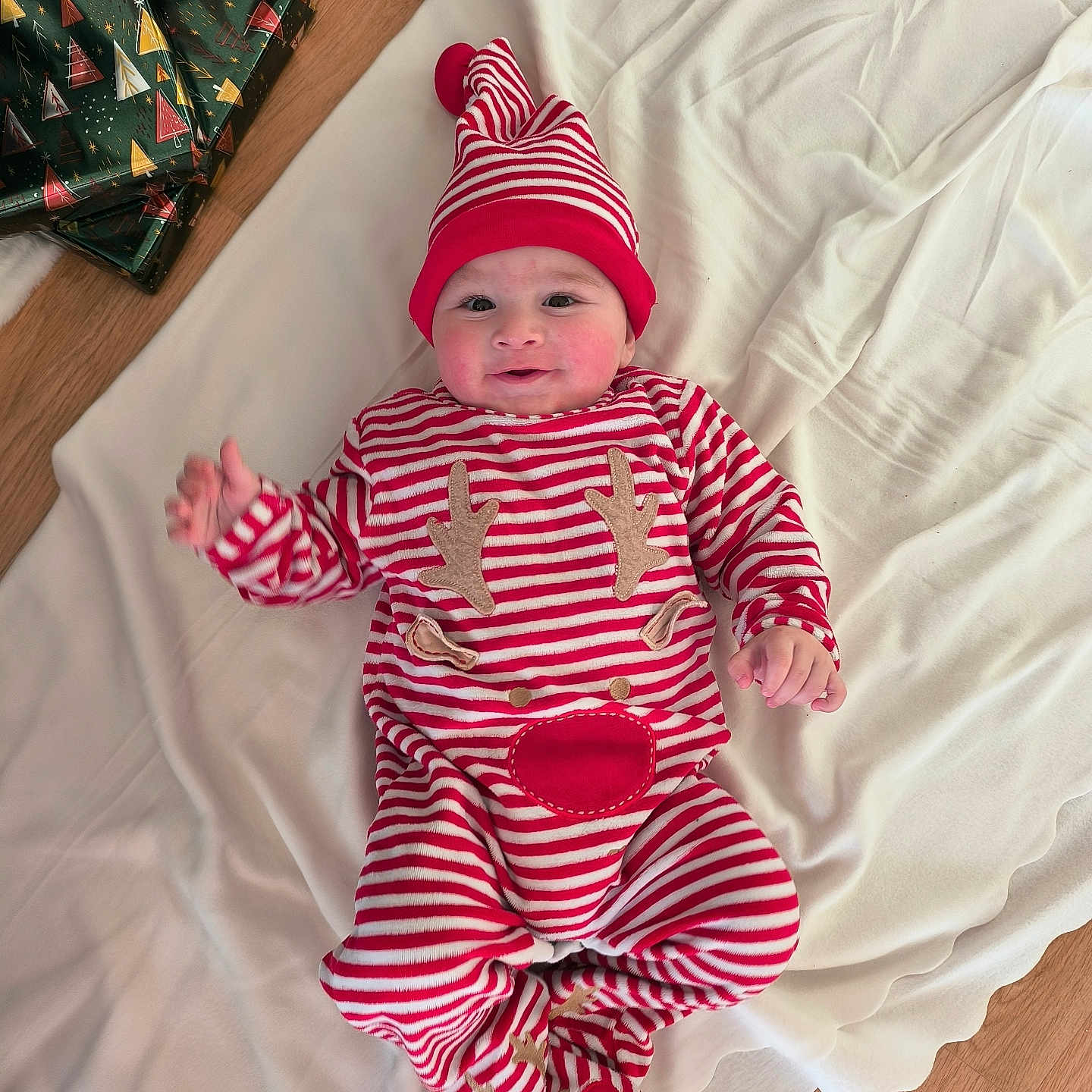 Aksel a rejoint le concours — aidez-le/la à gagner de superbes lots ! baby, blanket, child, christmas_theme, cozy, cute, festive, floor, happy, hat, holiday, indoors, infant, lying_down, red_and_white, reindeer_onesie, smiling, striped_clothing, wooden_floor, wrapped_presents
