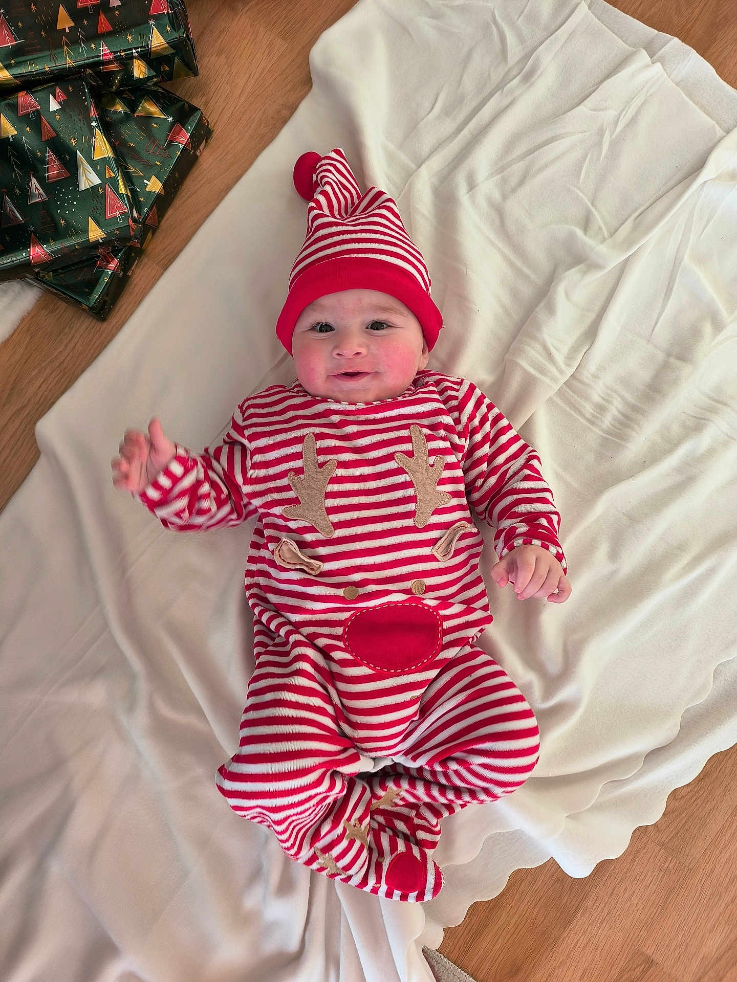 Aksel a rejoint le concours — aidez-le/la à gagner de superbes lots ! baby, infant, child, striped_clothing, red_and_white, reindeer_onesie, hat, blanket, floor, wrapped_presents, christmas_theme, smiling, cute, indoors, wooden_floor, holiday, festive, lying_down, cozy, happy