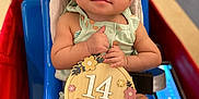 Amira is registered to the contest to win money with this photo: baby, child, infant, sitting, blue_chair, headband, green_headband, wooden_sign, 14_months, thumbs_up, cute, indoor, medical_tape, hospital_equipment, person, clothing, legs, face, smile, happy