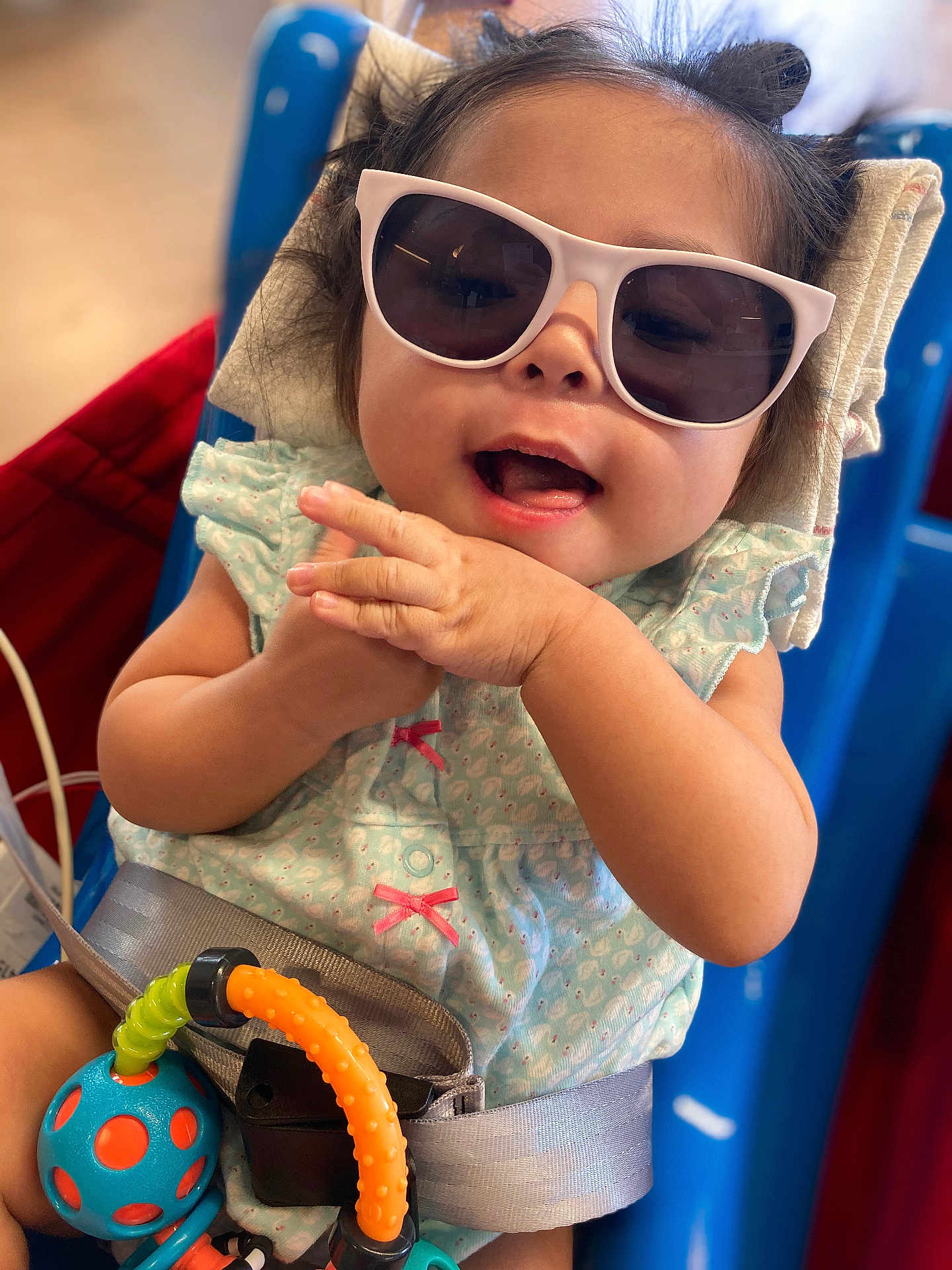 Amira is registered to the contest to win money with this photo: baby, child, sunglasses, smiling, toy, seatbelt, blue, orange, clothing, happy, cute, person, indoor, infant, hands, playful, accessory, closeup, seat, fun