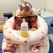Rosalina is registered to the contest to win money with this photo: baby, infant, headband, bottle, inflatable_seat, table, snacks, indoor, person, child, clothing, pink, white, face, feet, expression, home, decor, sitting, cute