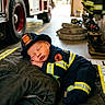 Christian joined the competition — help win amazing prizes! baby, child, sleeping, firefighter, uniform, fire_station, fire_engine, helmet, protective_gear, reflective_stripes, portrait, indoors, nap, cushions, equipment, boots, emergency_services, red, black, adorable