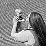 Kyler is registered to the contest to win money with this photo: baby, woman, brick_wall, smile, happy, holding, portrait, outdoor, black_and_white, person, long_hair, lace, clothing, joyful, face, child, interaction, cute, playful, toddler