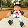 baby, baseball_cap, baseball_uniform, blanket, child, smiling, outdoor, dirt, sports_field, fence, greenery, person, cute, sitting, cap, happy, infant, clothing, portrait, playful