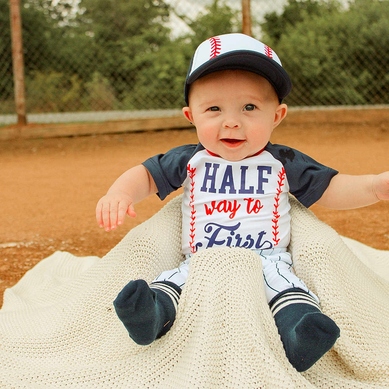 Kyler is registered to the contest to win money with this photo: baby, baseball_cap, baseball_uniform, blanket, child, smiling, outdoor, dirt, sports_field, fence, greenery, person, cute, sitting, cap, happy, infant, clothing, portrait, playful