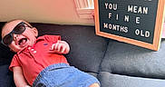 Kyler joined the competition — help win amazing prizes! baby, child, sunglasses, couch, window, letter_board, red_shirt, blue_shorts, smiling, happy, indoor, infant, person, casual_clothing, relaxing, sunlight, home, cute, funny_text, portrait