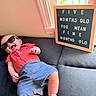baby, child, sunglasses, couch, window, letter_board, red_shirt, blue_shorts, smiling, happy, indoor, infant, person, casual_clothing, relaxing, sunlight, home, cute, funny_text, portrait