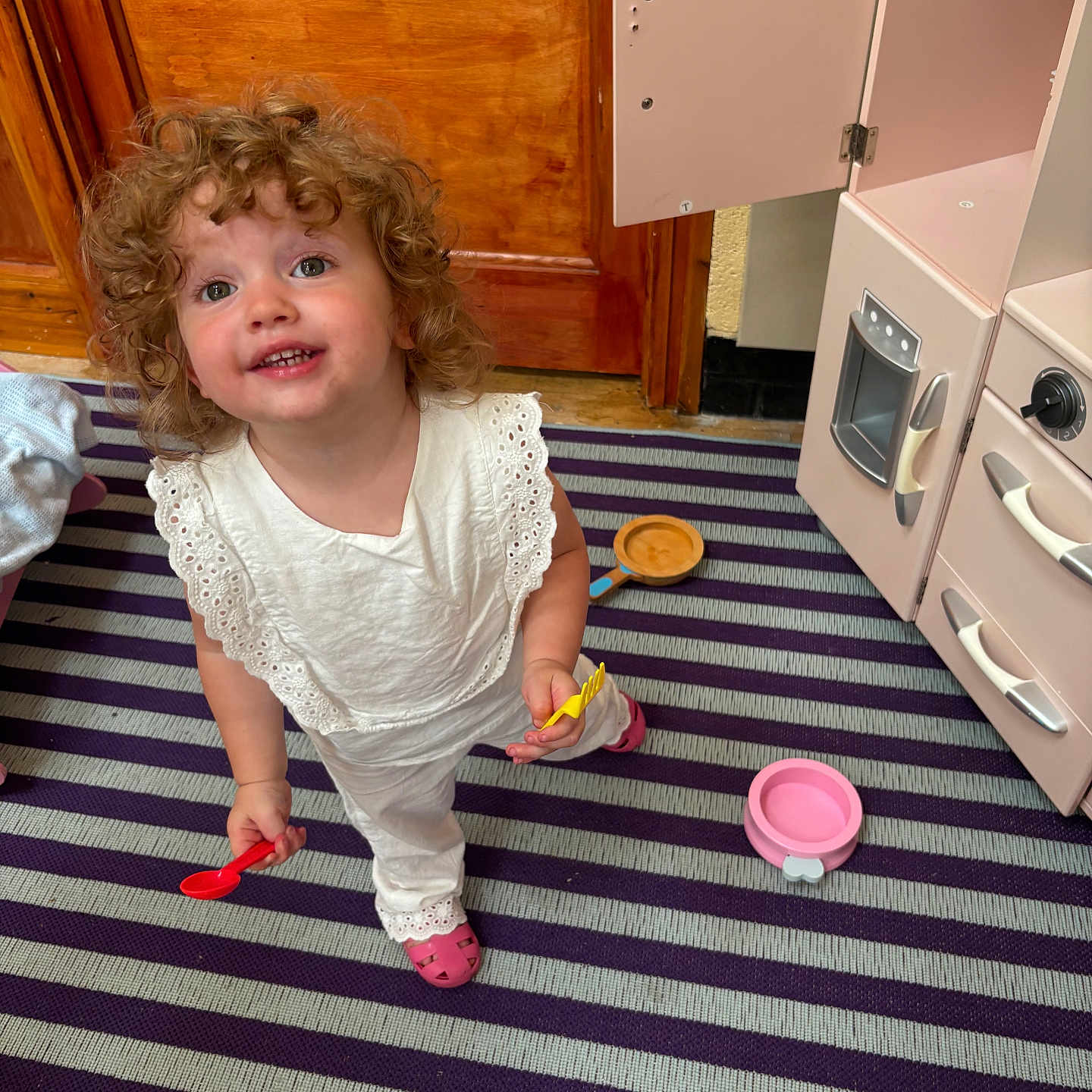 Harriet joined the competition — help win amazing prizes! child, curly_hair, cute, face, floor, happy, indoors, kitchen_toys, person, pink_shoes, play_kitchen, playtime, smiling, standing, striped_rug, toddler, toy_utensils, white_clothing, wooden_door, young_child