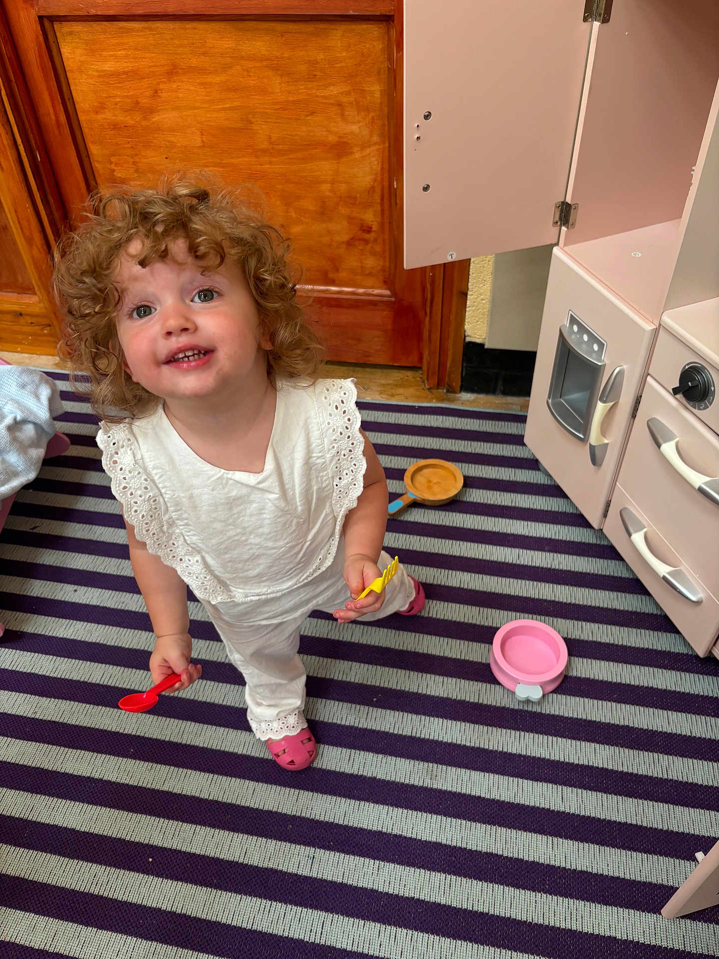 Harriet joined the competition — help win amazing prizes! toddler, child, curly_hair, smiling, white_clothing, pink_shoes, toy_utensils, striped_rug, wooden_door, play_kitchen, indoors, happy, cute, standing, floor, young_child, playtime, kitchen_toys, face, person