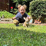 Harriet joined the competition — help win amazing prizes! toddler, child, garden, grass, plants, flowers, statues, rabbit_statues, greenery, sunlight, happy, curly_hair, outdoor, crouching, playful, decorations, nature, smiling, blue_shirt, black_pants