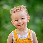 Mayron a rejoint le concours — aidez-le/la à gagner de superbes lots ! toddler, child, curly_hair, smiling, orange_overalls, white_shirt, outdoor, green_background, portrait, happy, cute, person, young_child, innocence, natural_light, close_up, face, eyes, skin, baby