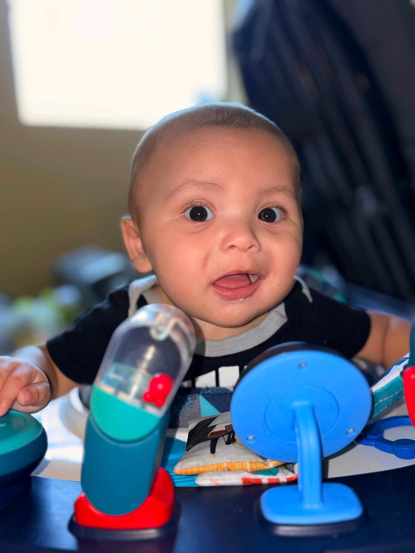 Kyire is registered to the contest to win money with this photo: baby, black_clothing, blue_toy, bright, cheek, child, closeup, colorful, curious, expression, face, happy, indoor, infant, plastic_toy, play, red_toy, smile, tongue, toy