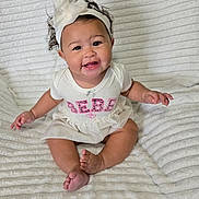 Anika is registered to the contest to win money with this photo: baby, child, smiling, headband, white_clothing, sitting, blanket, cute, happy, indoors, portrait, infant, toddler, pink_text, bow, soft_texture, human, young_child, playful, fun
