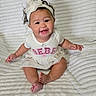 baby, child, smiling, headband, white_clothing, sitting, blanket, cute, happy, indoors, portrait, infant, toddler, pink_text, bow, soft_texture, human, young_child, playful, fun