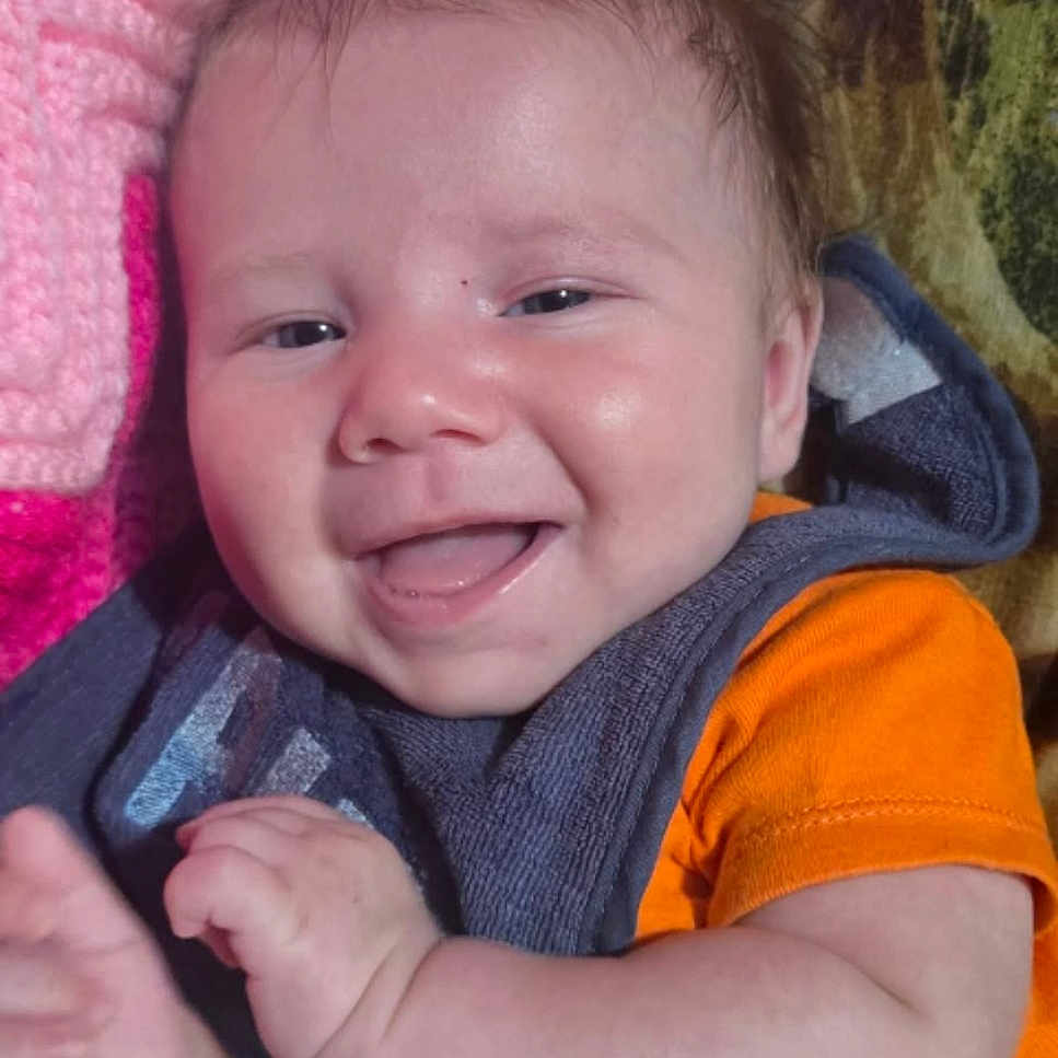 Brigham joined the competition — help win amazing prizes! baby, bib, child, closeup, crochet, cute, expression, face, hair, hand, happy, indoors, infant, lying_down, orange_shirt, person, pink_blanket, portrait, skin, smiling