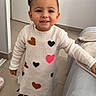 child, toddler, indoor, smiling, sweater_dress, hearts, sneakers, floor_tiles, door, blanket, couch, leaning, happy, person, portrait, casual_clothing, hair_buns, young_child, cute, playful