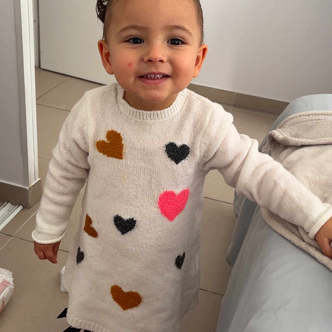 Kamila a rejoint le concours — aidez-le/la à gagner de superbes lots ! child, toddler, indoor, smiling, sweater_dress, hearts, sneakers, floor_tiles, door, blanket, couch, leaning, happy, person, portrait, casual_clothing, hair_buns, young_child, cute, playful