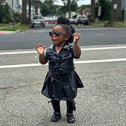 Charis is registered to the contest to win money with this photo: toddler, child, outdoor, street, sunglasses, black_clothing, leather, standing, fashion, confident, shoes, sock, hair, curly_hair, urban, sidewalk, road, trees, daytime, person