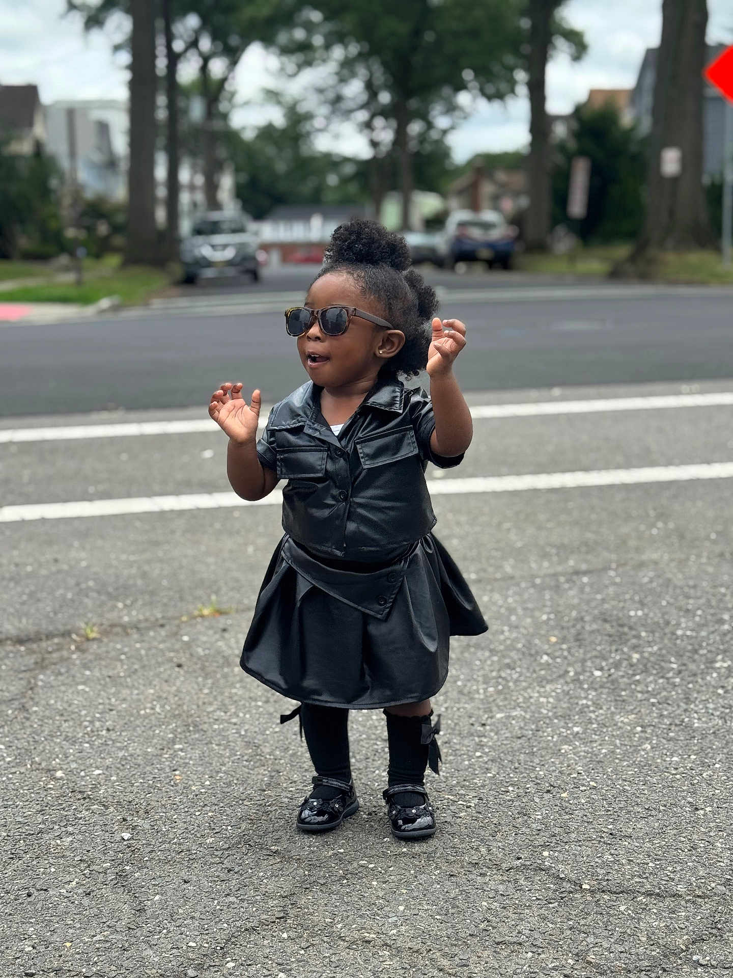 Charis is registered to the contest to win money with this photo: toddler, child, outdoor, street, sunglasses, black_clothing, leather, standing, fashion, confident, shoes, sock, hair, curly_hair, urban, sidewalk, road, trees, daytime, person