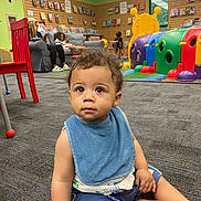 Cavean is registered to the contest to win money with this photo: toddler, child, playroom, bib, carpet, red_chair, toy, couch, person, indoor, curly_hair, shorts, sitting, colorful, window, books, shelf, floor, relaxing, casual