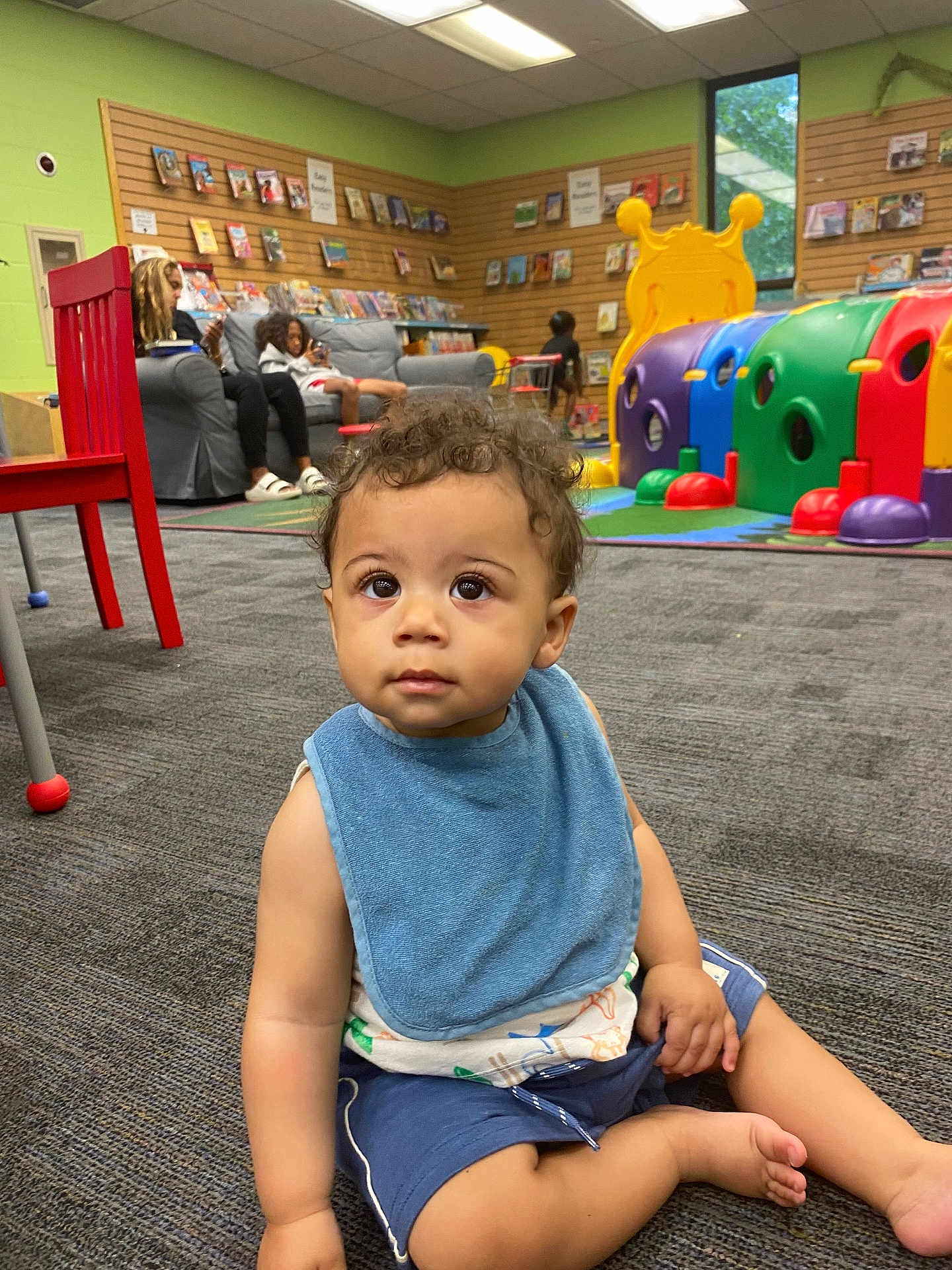 Cavean is registered to the contest to win money with this photo: toddler, child, playroom, bib, carpet, red_chair, toy, couch, person, indoor, curly_hair, shorts, sitting, colorful, window, books, shelf, floor, relaxing, casual
