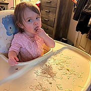 Tess is registered to the contest to win money with this photo: toddler, child, high_chair, spaghetti, messy, pink_onesie, finger_in_mouth, indoor, furniture, cabinet, clothing, tray, meal_time, food, cute, mess, portrait, baby, feeding, room