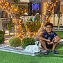 Ayser participe au concours pour gagner de l'argent avec cette photo : child, boy, smiling, grass, artificial_grass, tree, fairy_lights, dog_statue, outdoor, night, plants, bush, cactus, casual_clothing, shorts, sandals, watch, people, indoor, television