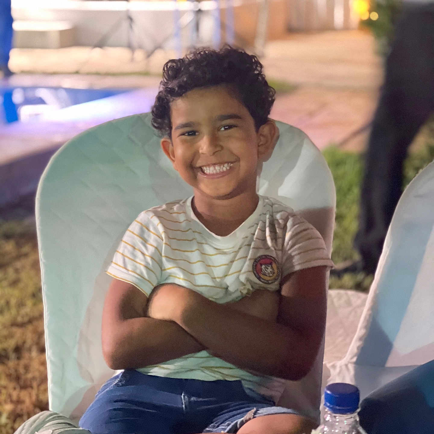 Ayser a rejoint le concours — aidez-le/la à gagner de superbes lots ! background_blur, bottle, boy, child, crossed_arms, curly_hair, flowers, grass, happy, night, outdoor, portrait, seated, shorts, smiling, sneakers, socks, striped_shirt, table, white_chair