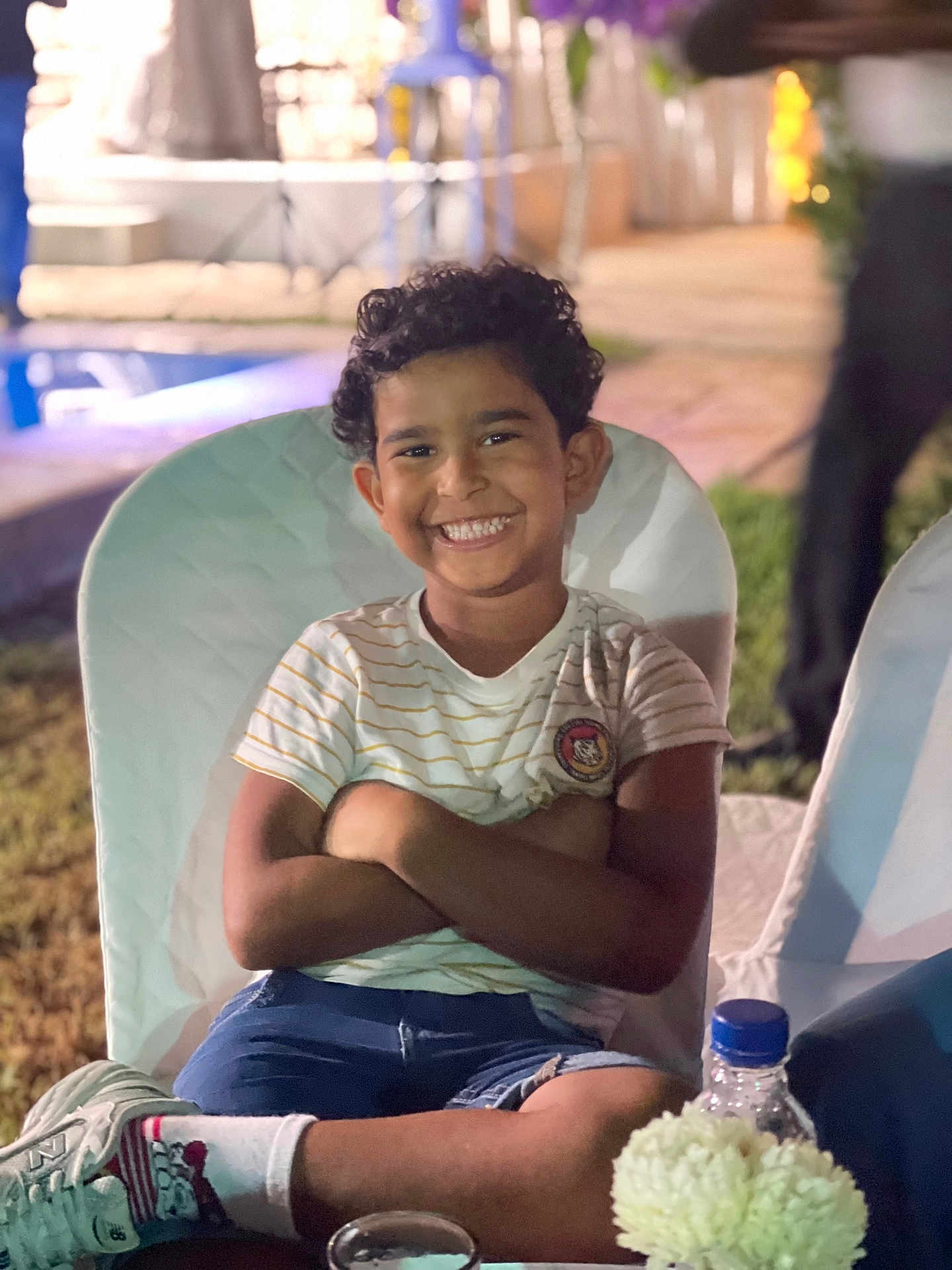 Ayser a rejoint le concours — aidez-le/la à gagner de superbes lots ! boy, child, smiling, curly_hair, white_chair, striped_shirt, shorts, socks, sneakers, outdoor, night, grass, bottle, flowers, table, happy, portrait, crossed_arms, seated, background_blur