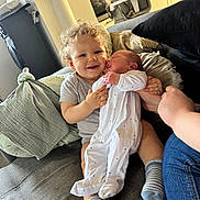 Gabyn a rejoint le concours — aidez-le/la à gagner de superbes lots ! adult_hand, baby, casual_clothing, child, close_up, couch, curly_hair, cushion, family, happy, holding, indoor, infant, living_room, newborn, onesie, person, smile, socks, toddler