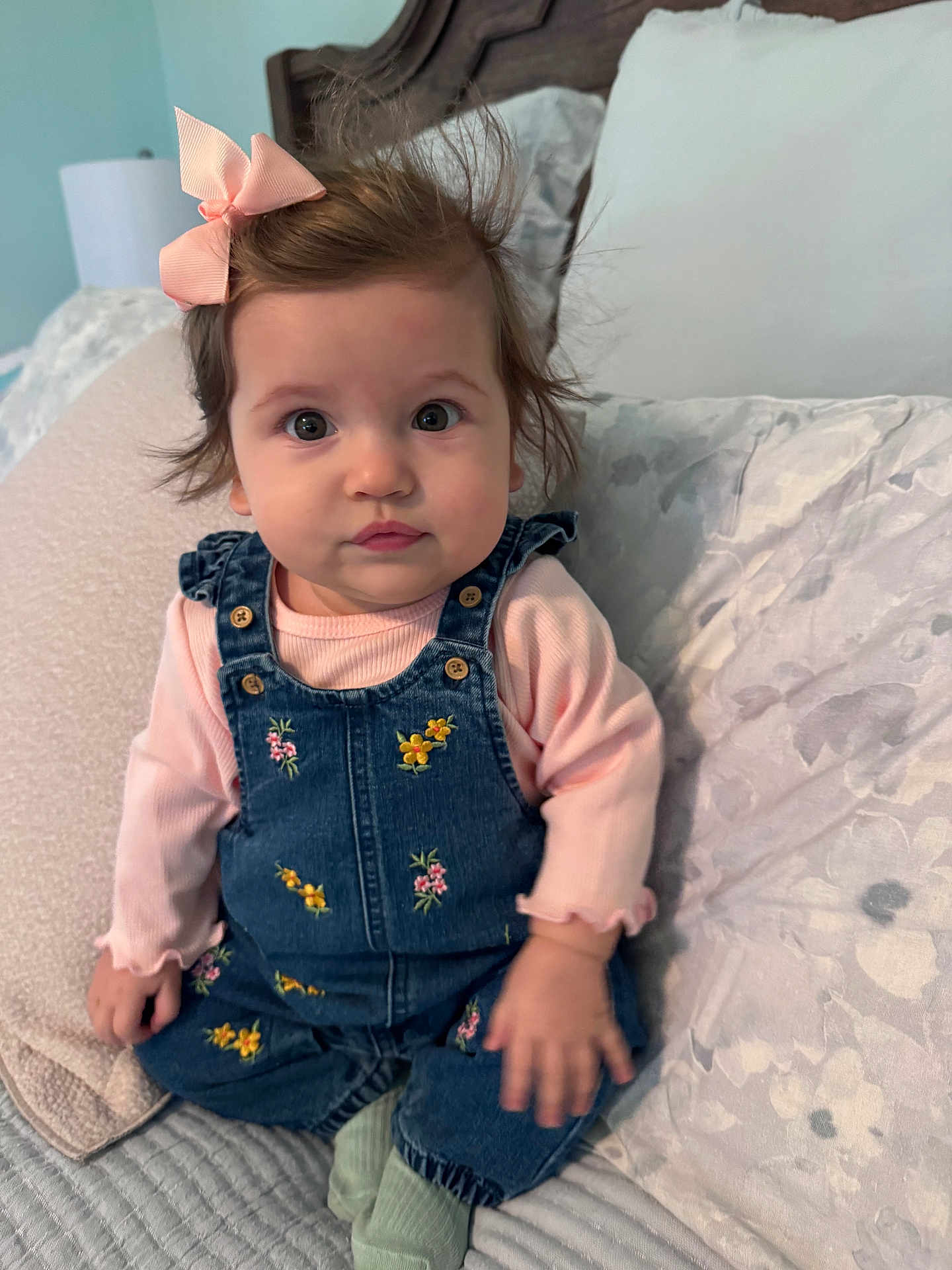 Virginia is registered to the contest to win money with this photo: baby, child, denim_overalls, pink_bow, long_sleeve_shirt, sitting, bed, pillow, blanket, indoor, curly_hair, cute, portrait, young_child, expression, cozy, soft_lighting, home, person, clothing