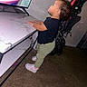 toddler, child, standing, indoors, television, watching_tv, stroller, table, marble_surface, pink_shoes, green_pants, black_shirt, curly_hair, profile, home, living_room, carpet, furniture, screen, portrait
