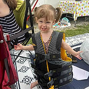 Gabriella is registered to the contest to win money with this photo: toddler, child, life_jacket, outdoor, camping, rug, pigtails, barefoot, smiling, person, adult, chair, table, plastic_toy, footwear, grass, patterned_textile, summer, family, fun