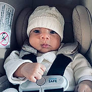 Adrián is registered to the contest to win money with this photo: baby, child, car_seat, white_hat, knitwear, baby_clothing, safety_harness, serious_face, infant, closeup, indoors, seat_belt, cute, person, headwear, small_hand, infant_face, child_safety, car_travel, protection