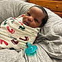 Noble is registered to the contest to win money with this photo: baby, infant, swaddle, blanket, footprint_pattern, pacifier, cushion, gray_fabric, wooden_dresser, indoor, portrait, face, curious, resting, comfort, newborn, child, sleepy, soft, cozy