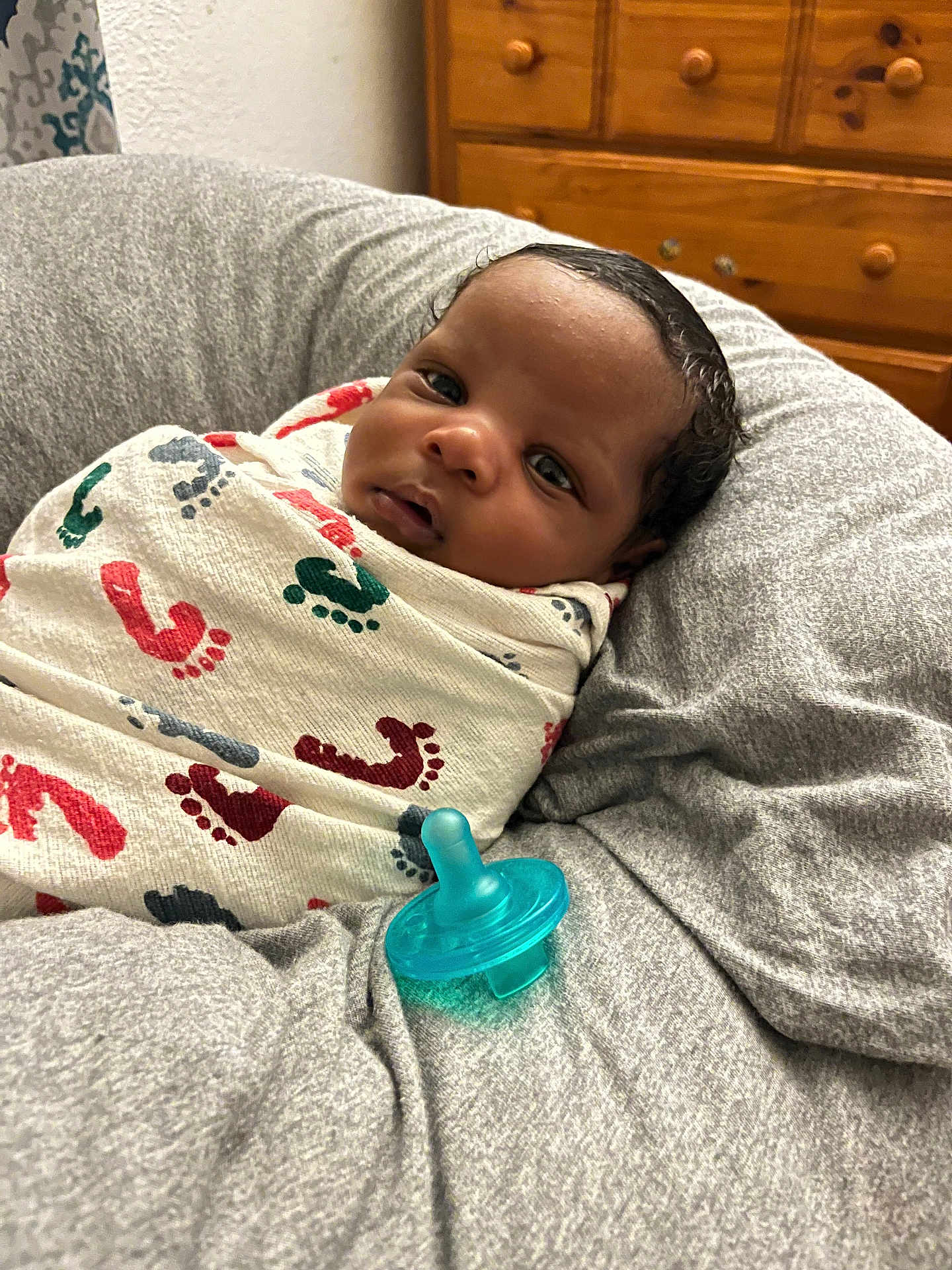 Noble is registered to the contest to win money with this photo: baby, infant, swaddle, blanket, footprint_pattern, pacifier, cushion, gray_fabric, wooden_dresser, indoor, portrait, face, curious, resting, comfort, newborn, child, sleepy, soft, cozy