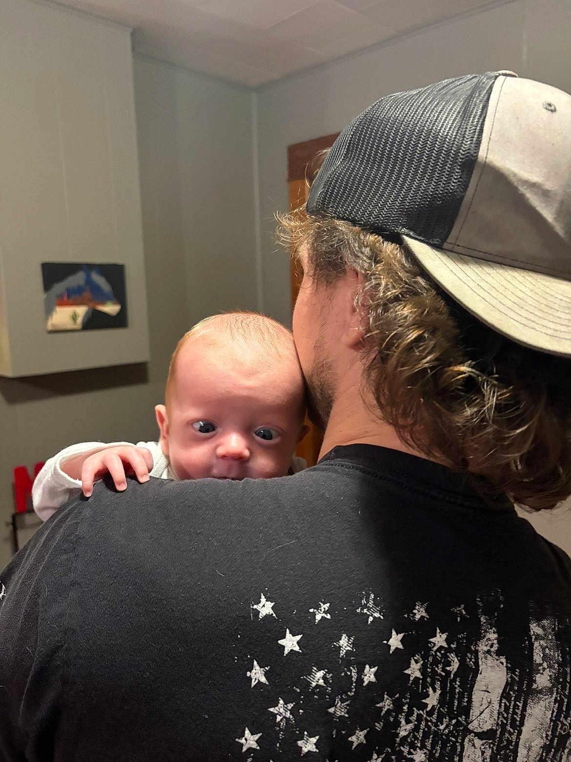 Liam Hughes is registered to the contest to win money with this photo: baby, person, indoor, hat, black_shirt, stars, curly_hair, shoulder, face, hand, expression, light, wall, painting, wooden_door, clothing, young_child, holding, head, closeup