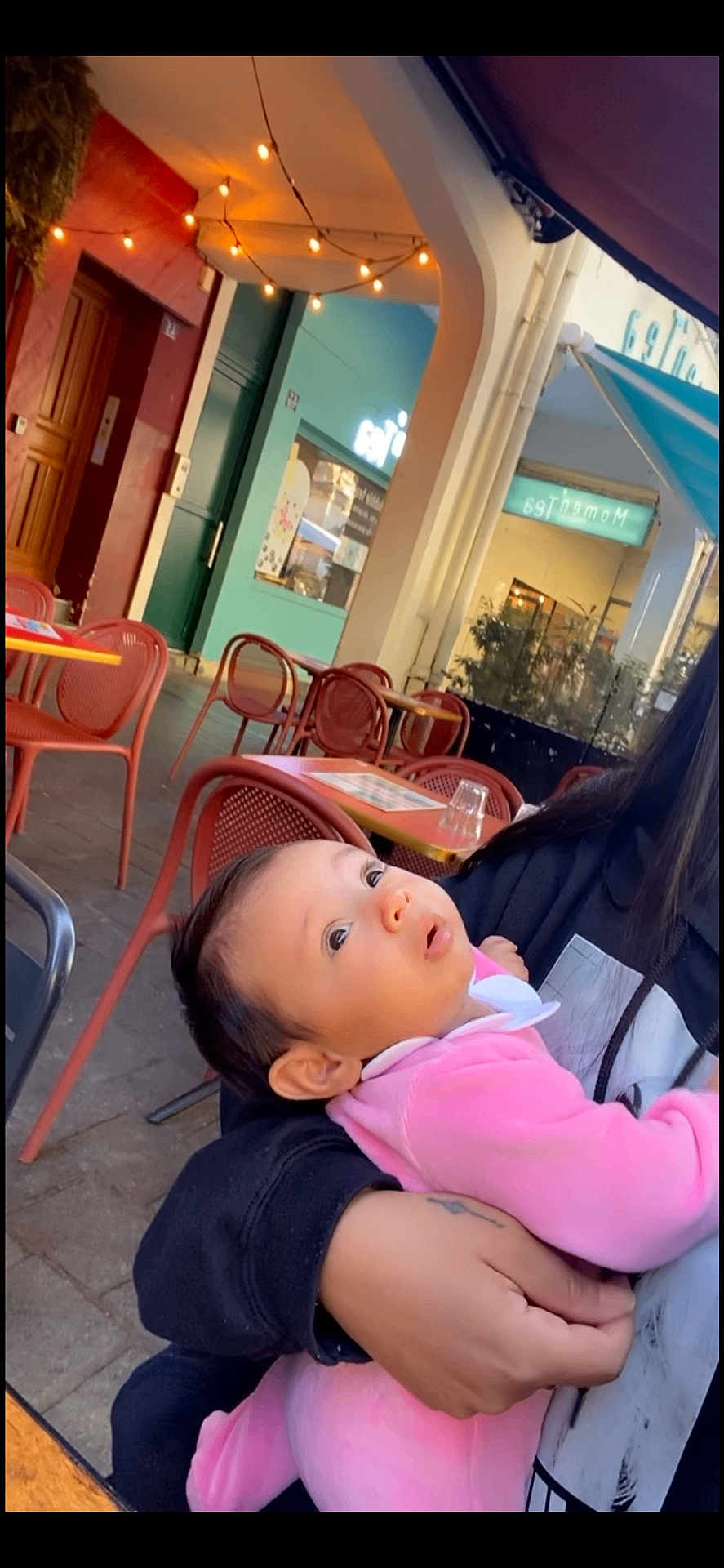 Julia a rejoint le concours — aidez-le/la à gagner de superbes lots ! baby, child, person, pink_clothing, holding, outdoor, cafe, chairs, table, string_lights, curious, tattoo, hoodie, face, portrait, seated, daylight, window, hand, expression