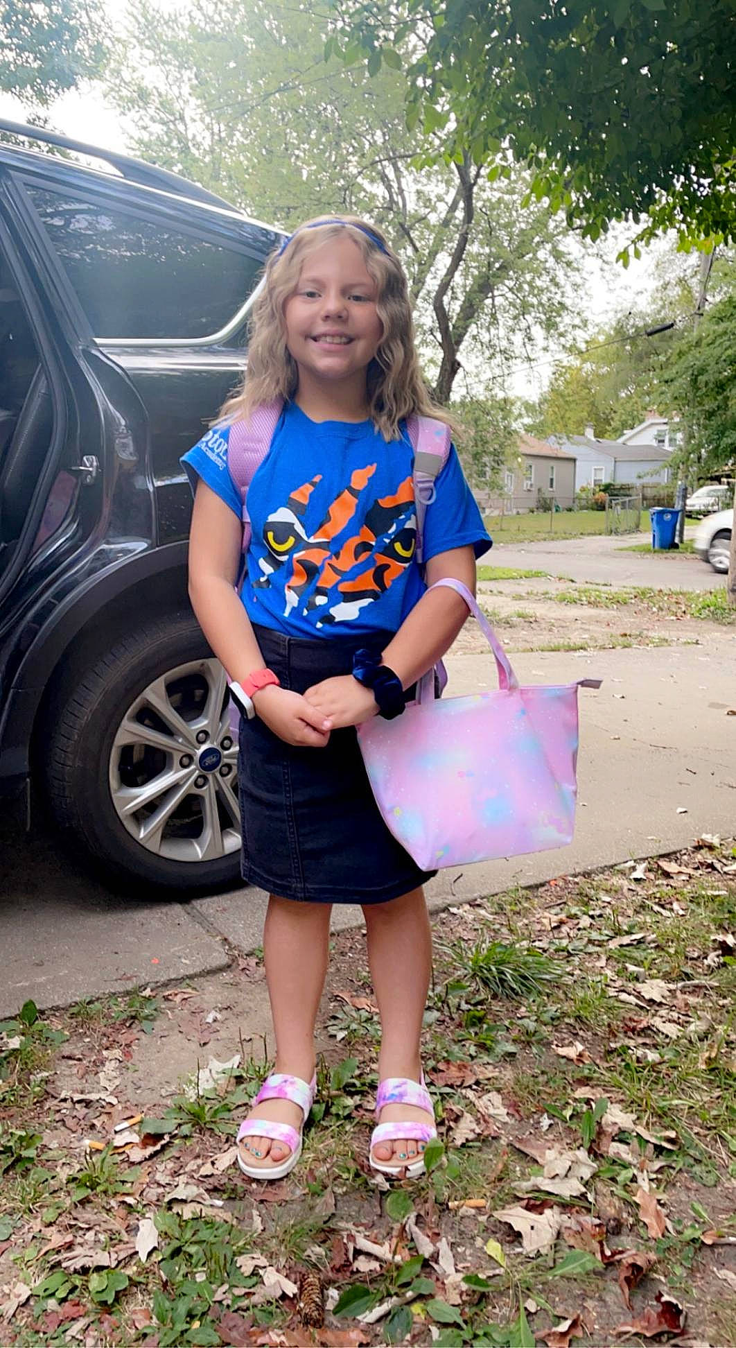 Evelina is registered to the contest to win money with this photo: automotive_exterior, blond, car, child, electric_blue, footwear, grass, joy, leisure, pattern, person, plant, recreation, shoe, smile, sneakers, t_shirt, tire, tree, vehicle_door