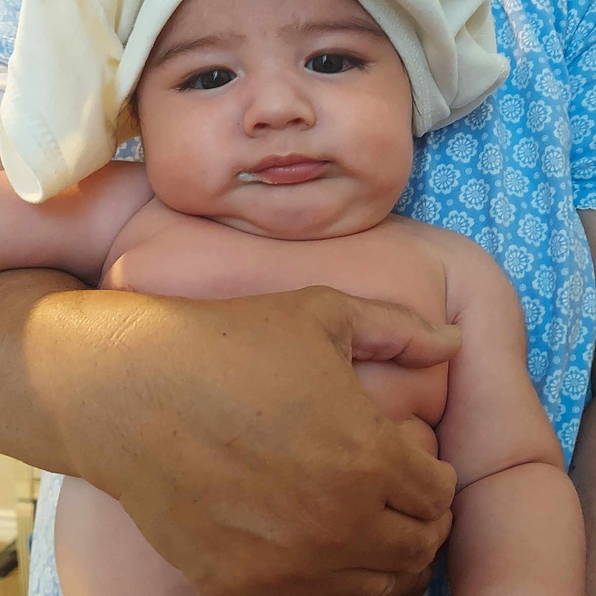 Tevita is registered to the contest to win money with this photo: baby, infant, towel, diaper, hand, adult, blue_clothing, floral_pattern, skin, face, expression, indoor, person, holding, cute, chubby, milk, portrait, child, closeup