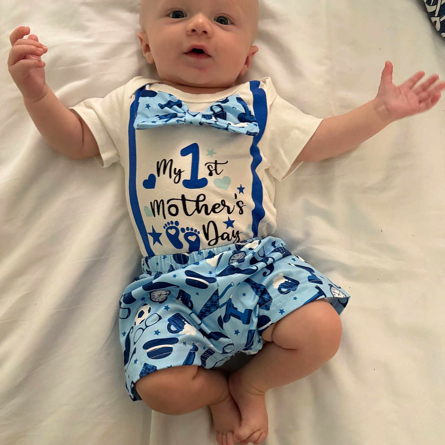 Kentley is registered to the contest to win money with this photo: arms_outstretched, baby, blue_bow_tie, blue_shorts, celebration, child, clothing, cute, face, first_mothers_day, footwear_none, happy, indoor, infant, lying_down, person, portrait, smiling, toddler, white_bed