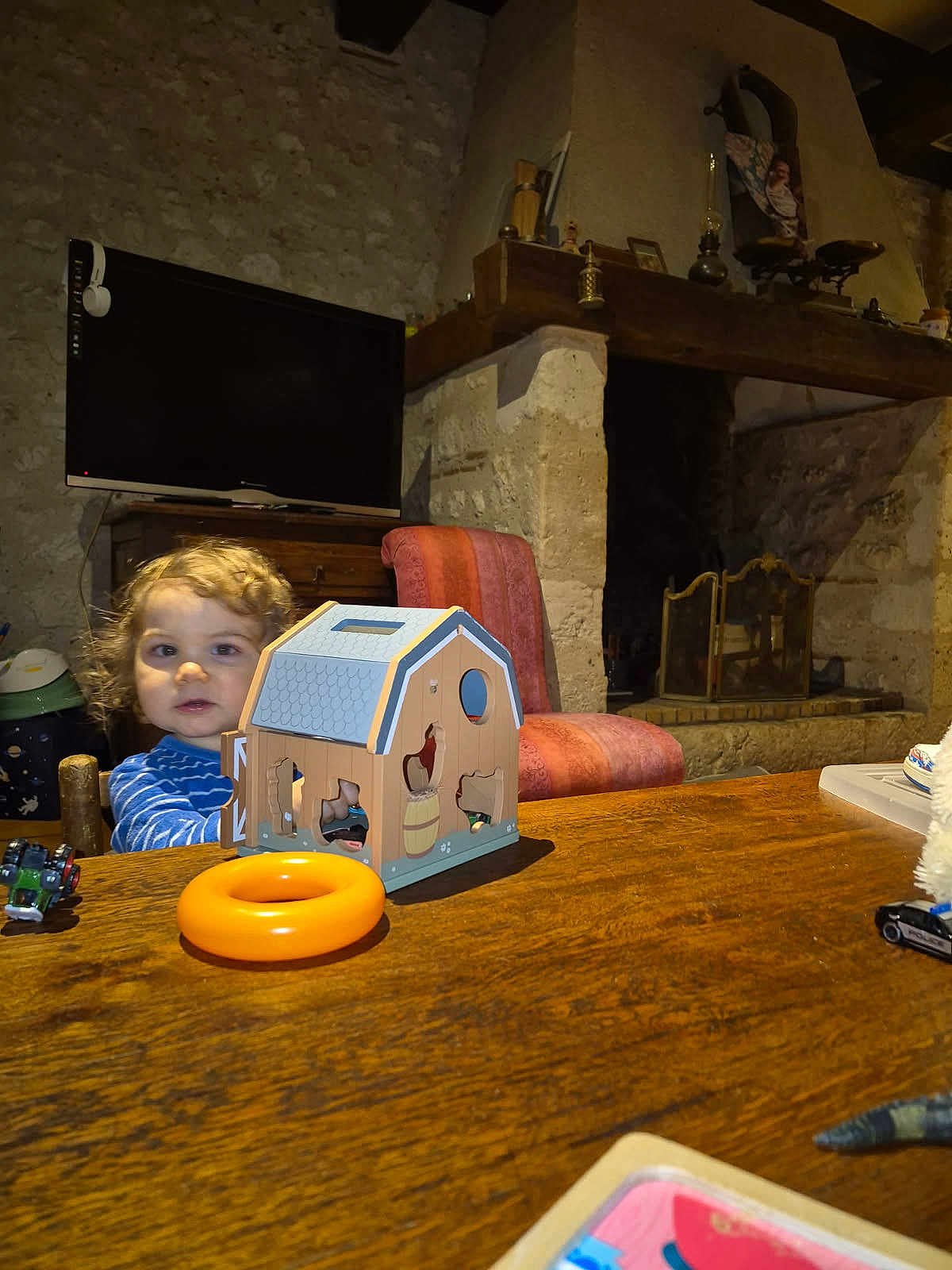Marcus participe au concours pour gagner de l'argent avec cette photo : toddler, child, toy, wooden_table, barn_puzzle, orange_ring, curly_hair, indoor, fireplace, stone_wall, chair, television, furniture, rustic, playtime, person, home, cozy, wood, curious