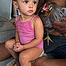 child, toddler, pink_tank_top, barefoot, sitting, chicken, adult_hand, floor, indoor, curly_hair, hair_ties, earrings, animal, pet, casual, expression, person, skin, holding, concrete_floor