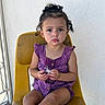 toddler, child, curly_hair, purple_clothing, yellow_chair, toy_spider, serious_expression, indoor, sitting, person, earrings, shorts, buttoned_top, light_skin, natural_light, plain_wall, texture, closeup, small_hands, curly_bun