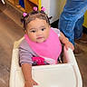 Armanii joined the competition — help win amazing prizes! baby, high_chair, pink_bib, hair_clips, child, indoor, wooden_floor, person, jeans, footwear, cute, smiling, face, hand, clothing, table, furniture, infant, seated, curious