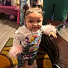 adult, balloon, carpet, child, clothing, colorful, decor, floor, fur_trim, hair_buns, hand, happy, indoor, jeans, jewelry, person, room, smile, table, toddler