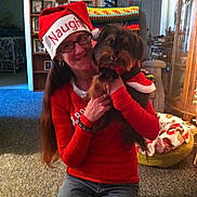 Elvis is registered to the contest to win money with this photo: woman, dog, indoor, holiday, christmas, hat, red_clothing, glasses, kneeling, carpet, furniture, blanket, dog_bed, crocheted_blanket, smiling, long_hair, casual, portrait, festive, home